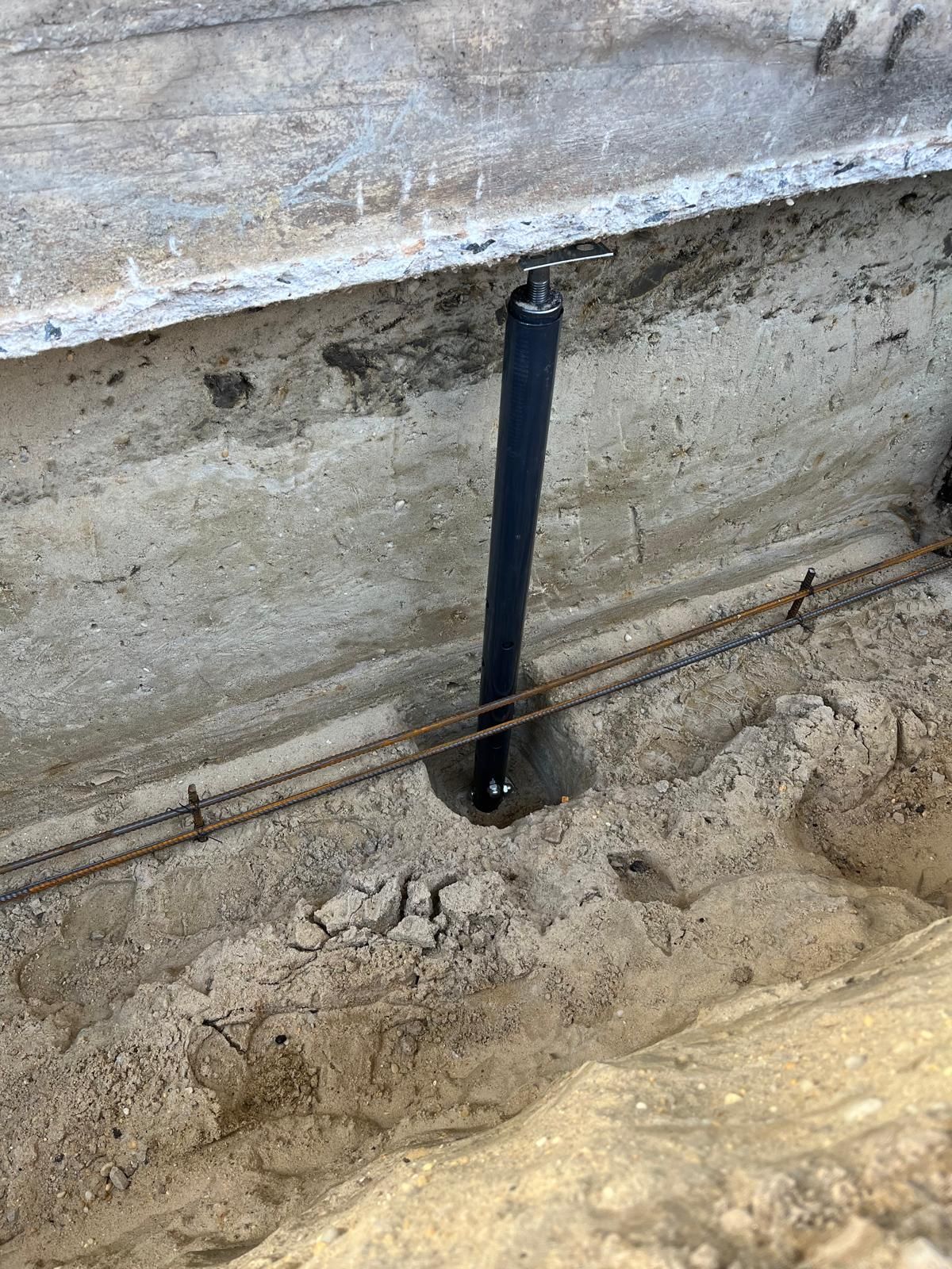 A vertical black pipe stands in a dirt trench next to a concrete wall with exposed steel reinforcement bars.