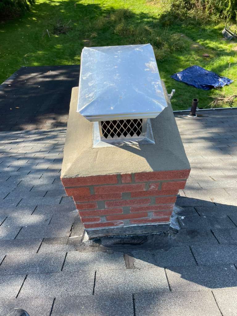 A red brick chimney with a new concrete crown and a metal cap, installed on a shingled residential roof.