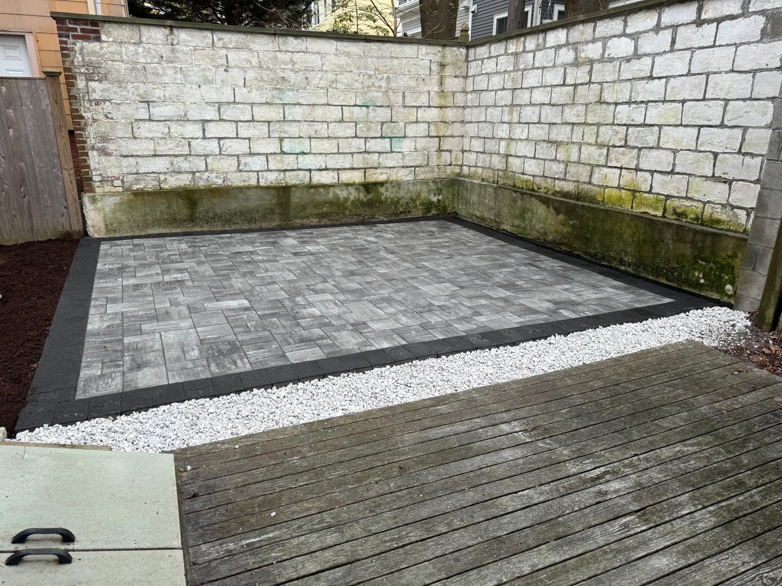 Small backyard patio with gray pavers, white gravel border, wooden deck, and mossy brick wall.