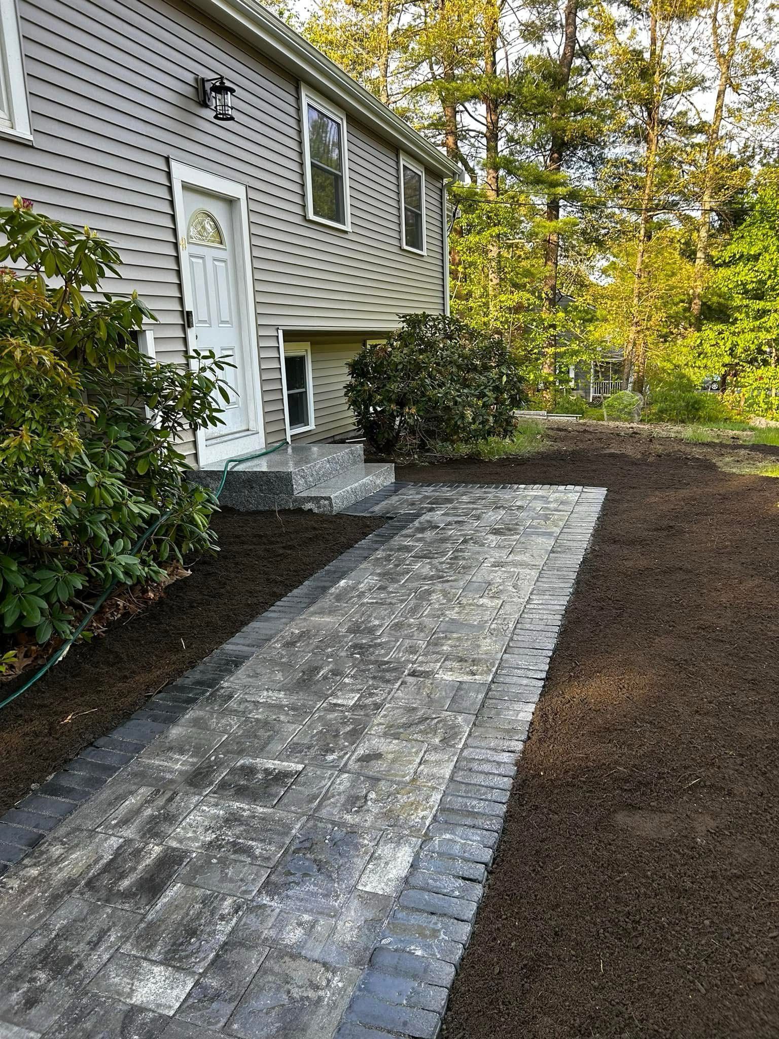 Stone walkway leading to a house entrance beside shrubs and a gravel path in a wooded yard