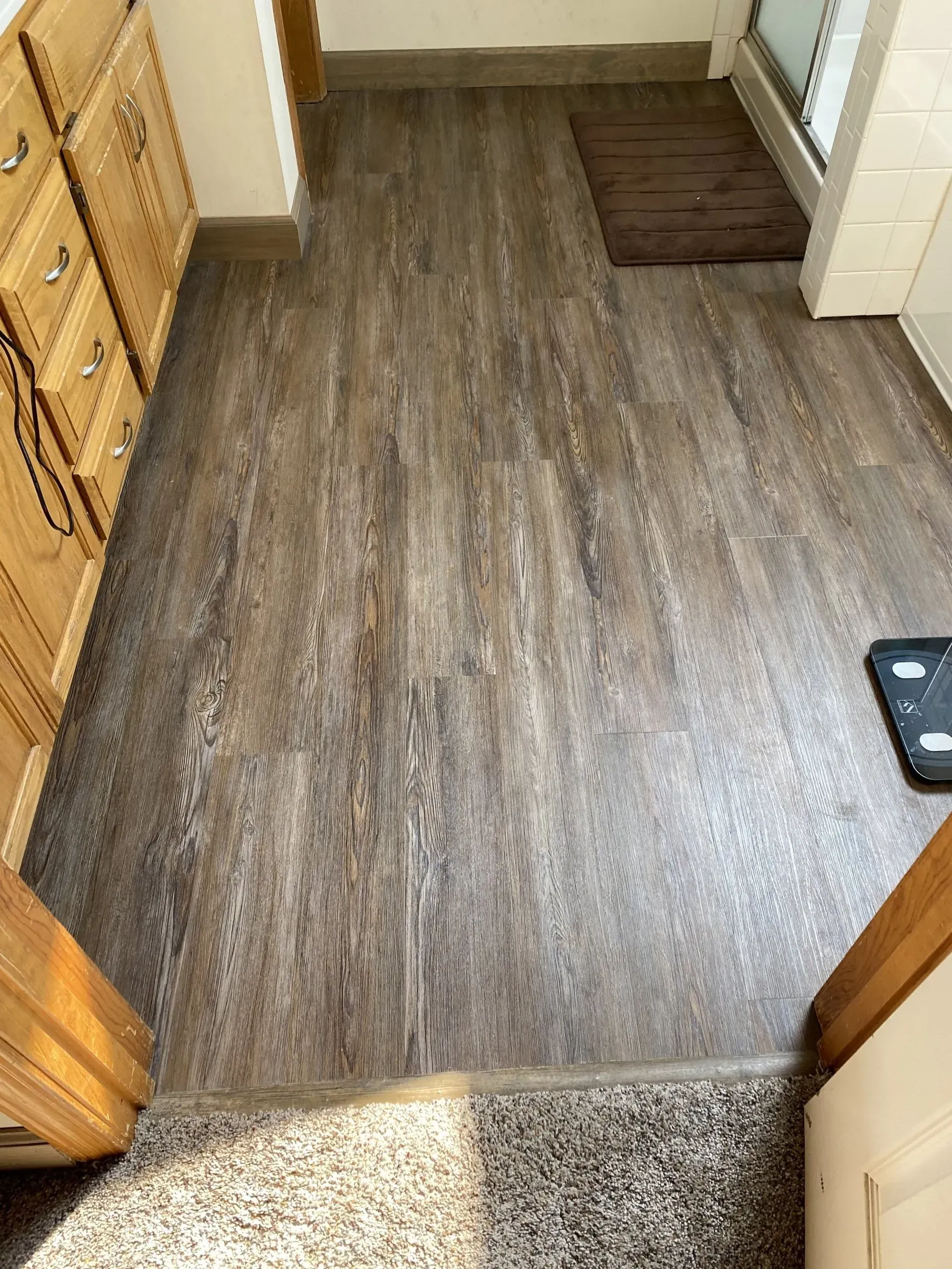 Bathroom with wood-look flooring. Light brown vanity on the left. Small brown rug and scale visible.