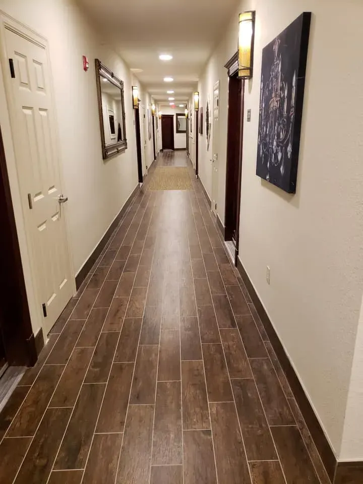 Long hallway with dark wood-look tile flooring, white walls, and dark wooden doors. Several light fixtures and artwork are present.