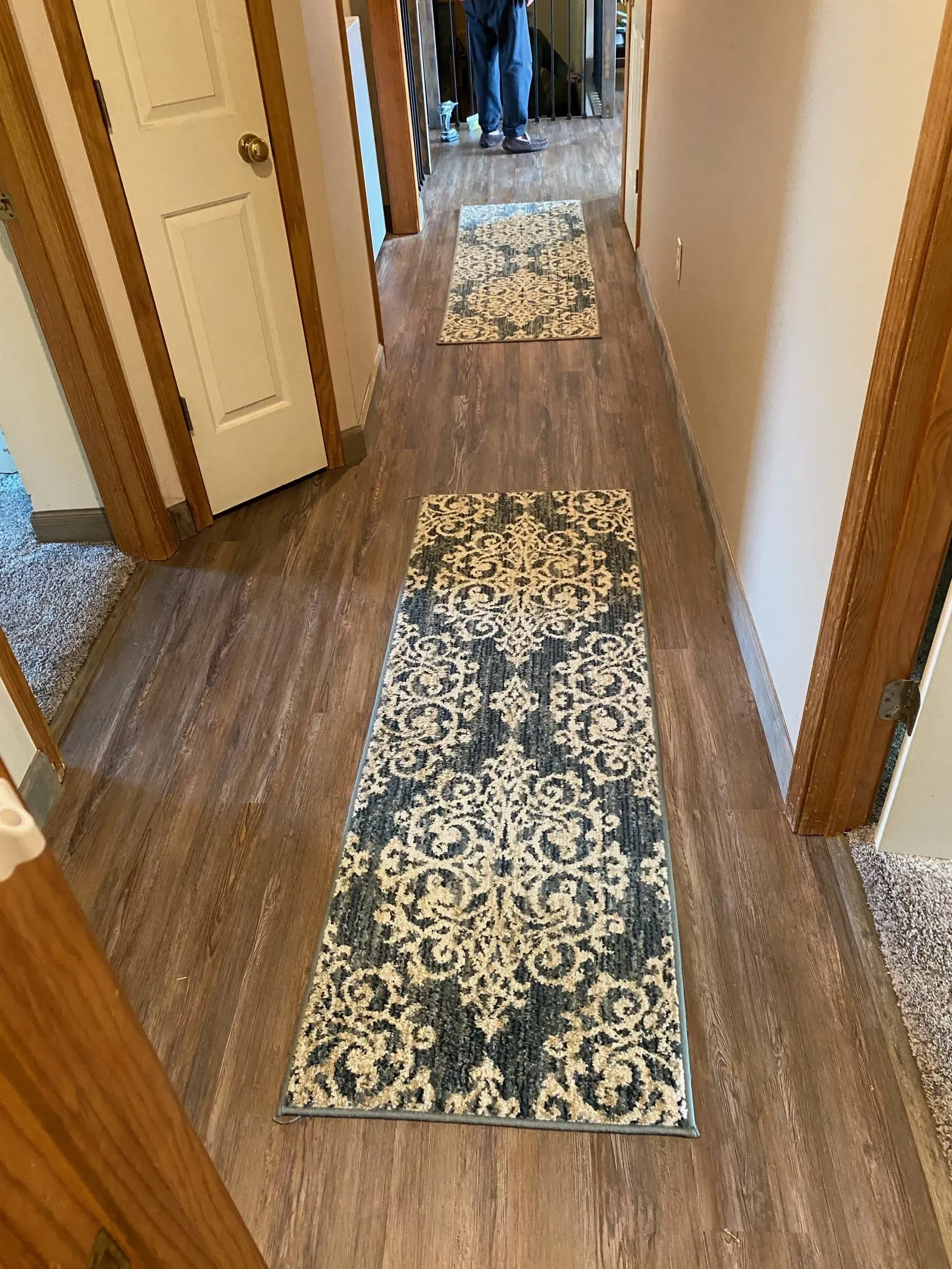 Hallway with wood-look flooring and two patterned runner rugs. A person stands at the end of the hall.