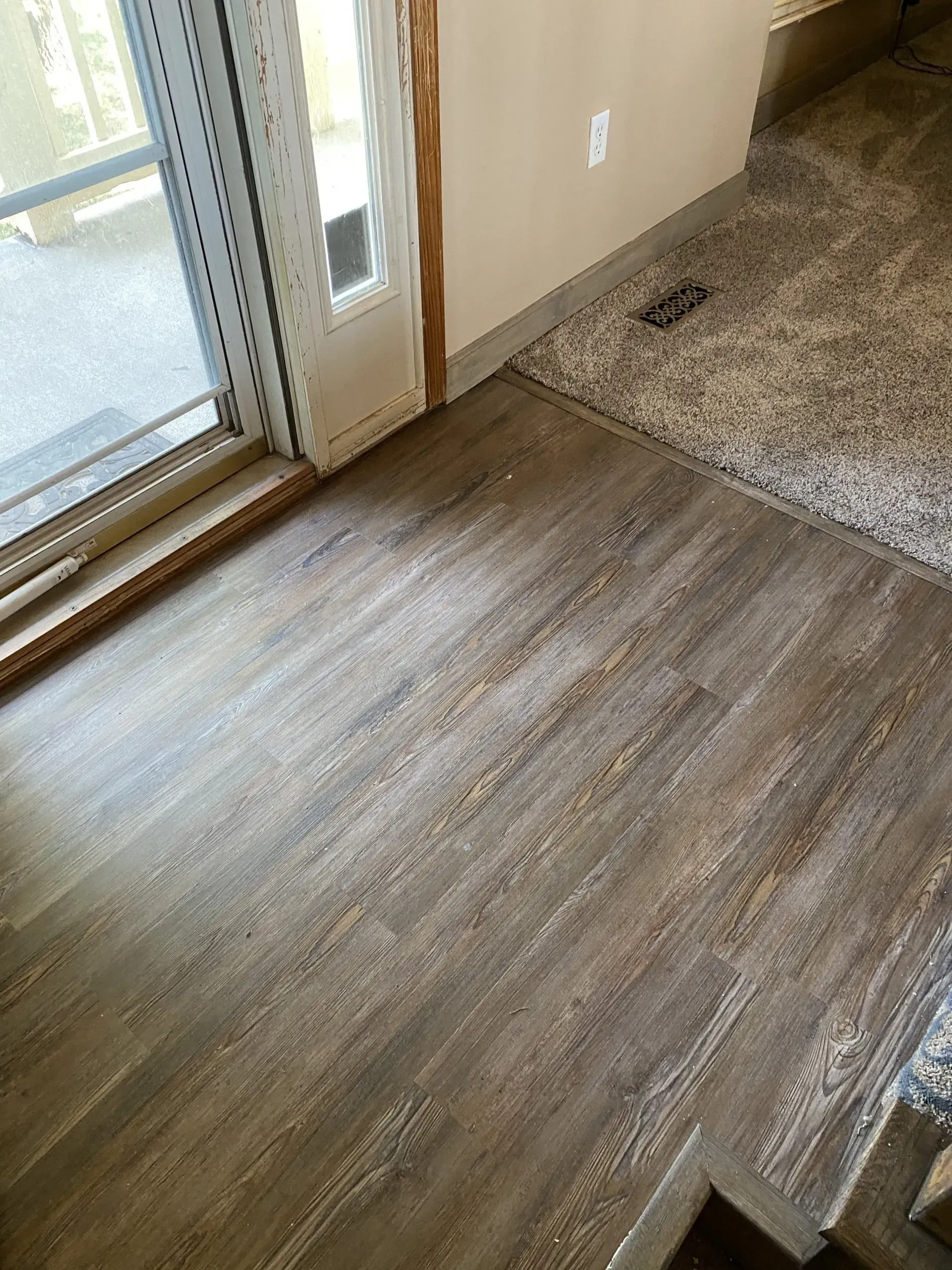 Wood-look flooring and carpet meet at a doorway. A sliding glass door and tan walls are also present.