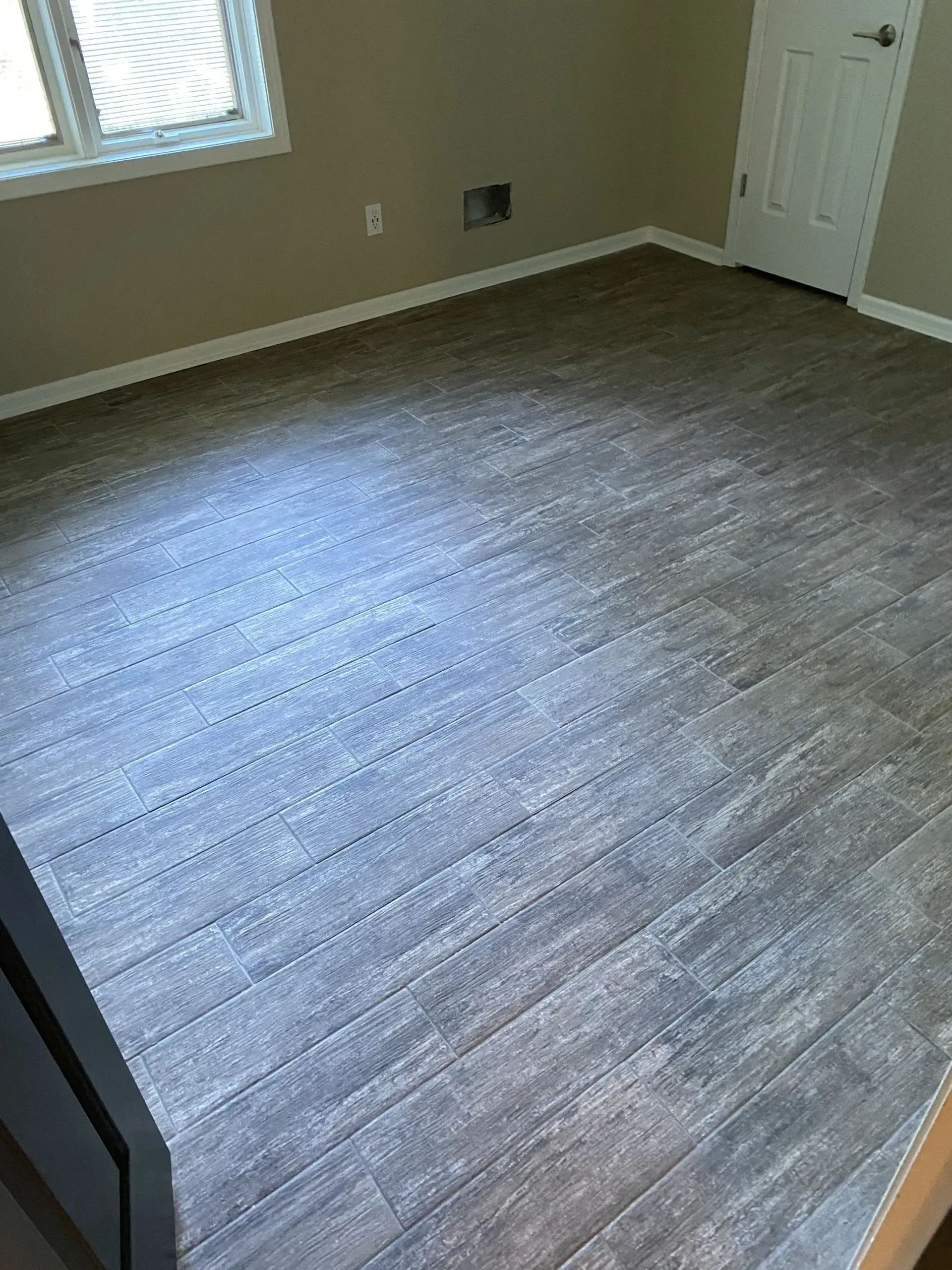 Room with wood-look flooring and a white door; a window and electrical outlet are visible on the wall.