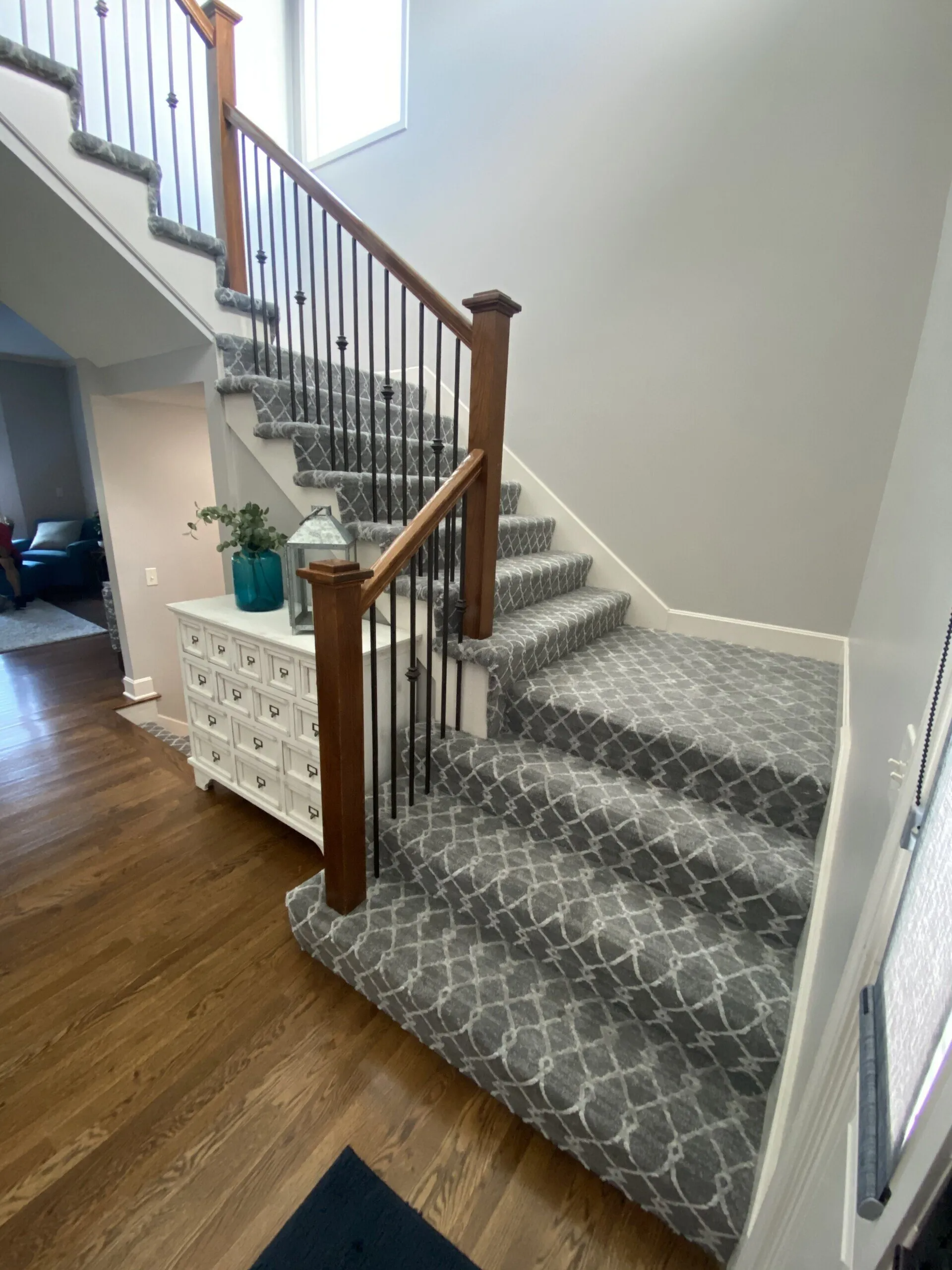 Staircase with patterned gray carpet and wooden railing. White dresser with decor at the base of the stairs.