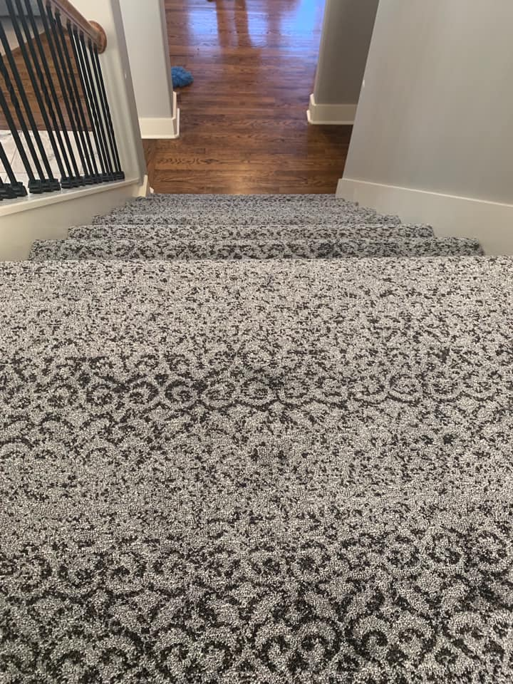 Staircase with patterned gray carpet leading down to hardwood flooring. Black railing on the left.