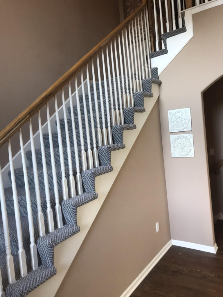 Staircase with white spindles, wooden handrail, and blue patterned carpet. Beige walls and white trim.