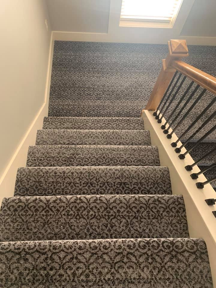 Staircase with gray patterned carpet, white risers, and a wooden railing with black spindles.