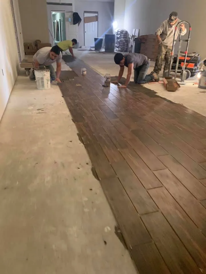 Construction workers installing wood-look tile on a concrete floor inside a room.