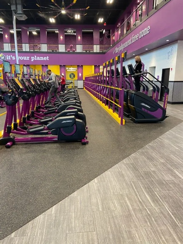 A gym interior with rows of elliptical machines and stair steppers; purple and yellow accents. A person uses a stepper.