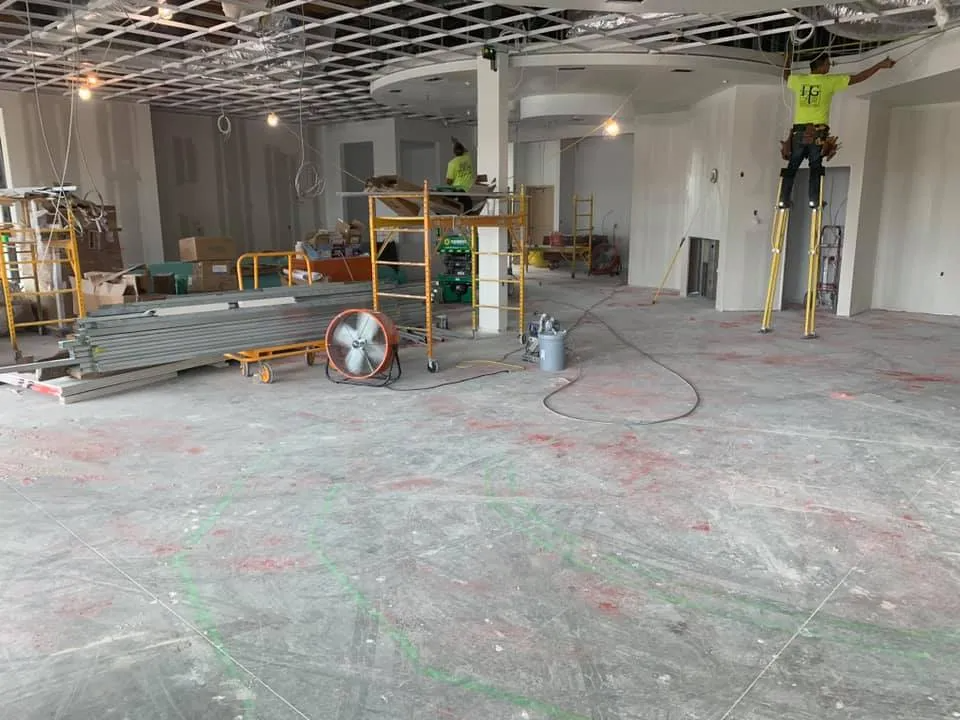 Construction workers inside a building. One on stilts working on the ceiling, others on scaffolding. Concrete floor.