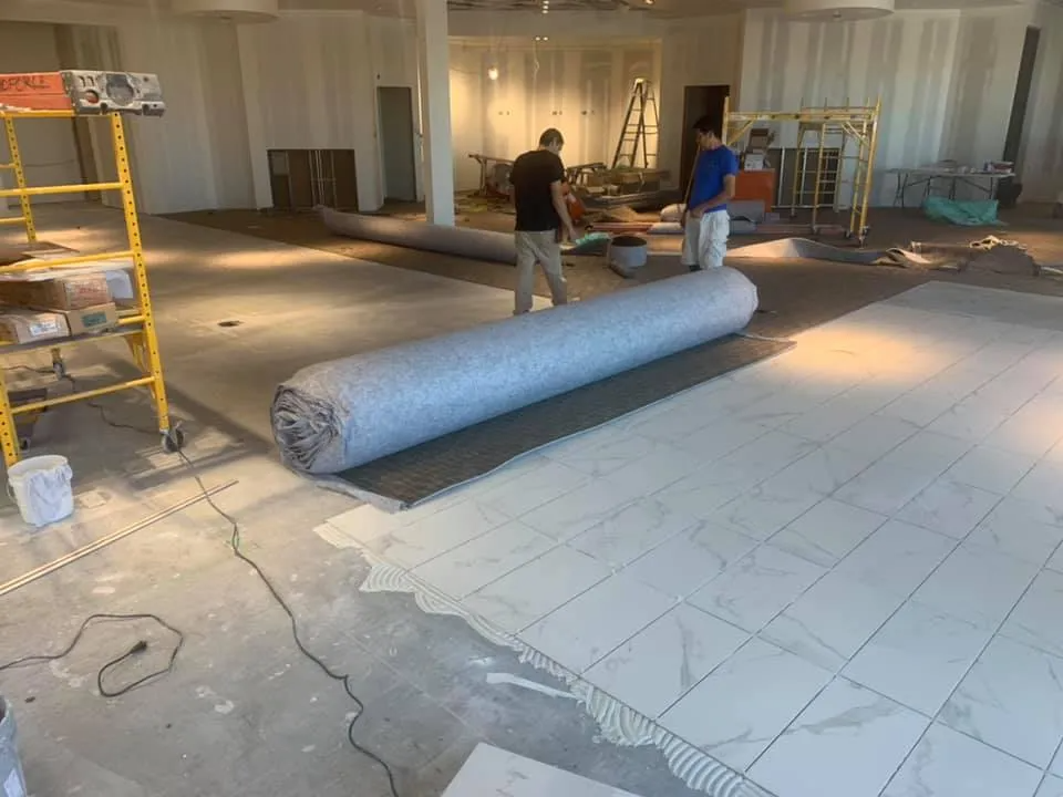 Workers installing flooring in an unfinished space; a rolled carpet, tiles, and scaffolding present.