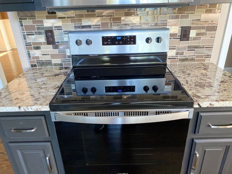 Stainless steel electric oven with black cooktop, in a kitchen with gray cabinets and stone backsplash.