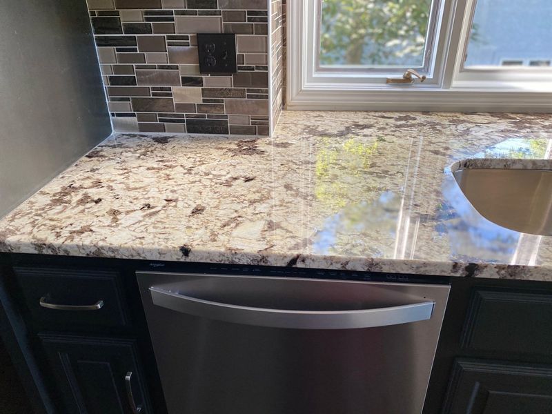 Kitchen countertop with granite and mosaic backsplash, stainless steel dishwasher under a window.