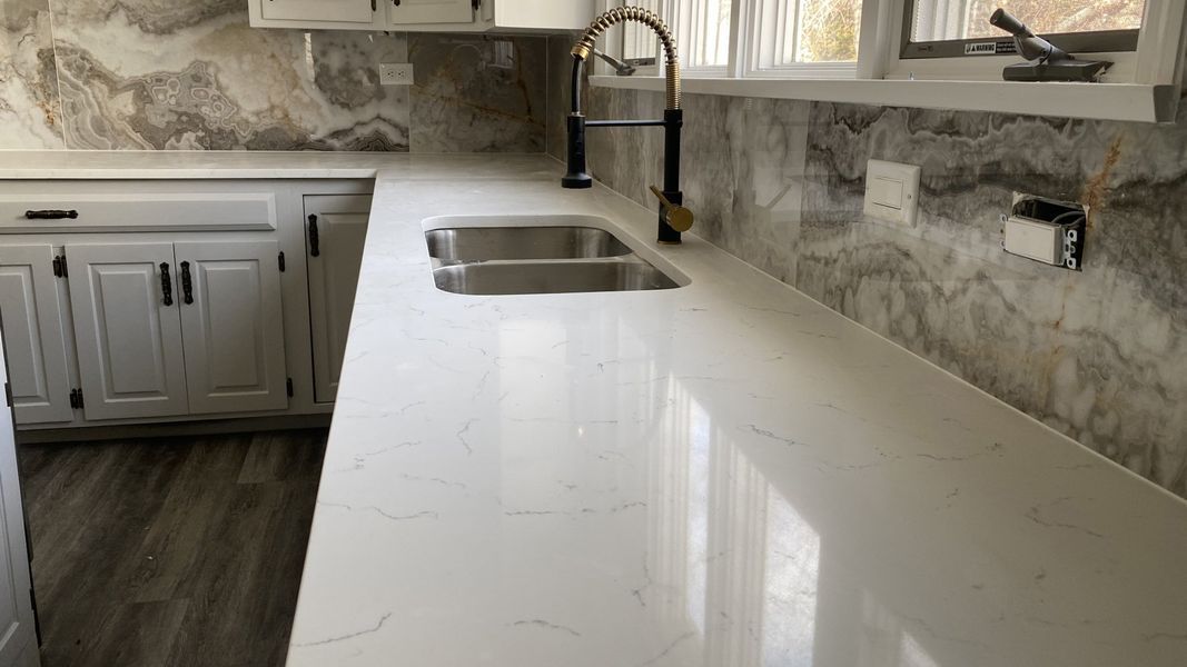 White kitchen countertop with stainless steel sink, gold faucet, and textured backsplash.
