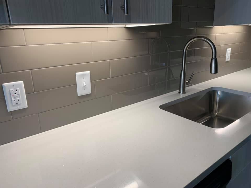 Kitchen counter with stainless steel sink, faucet, and backsplash, illuminated by cabinet lights.