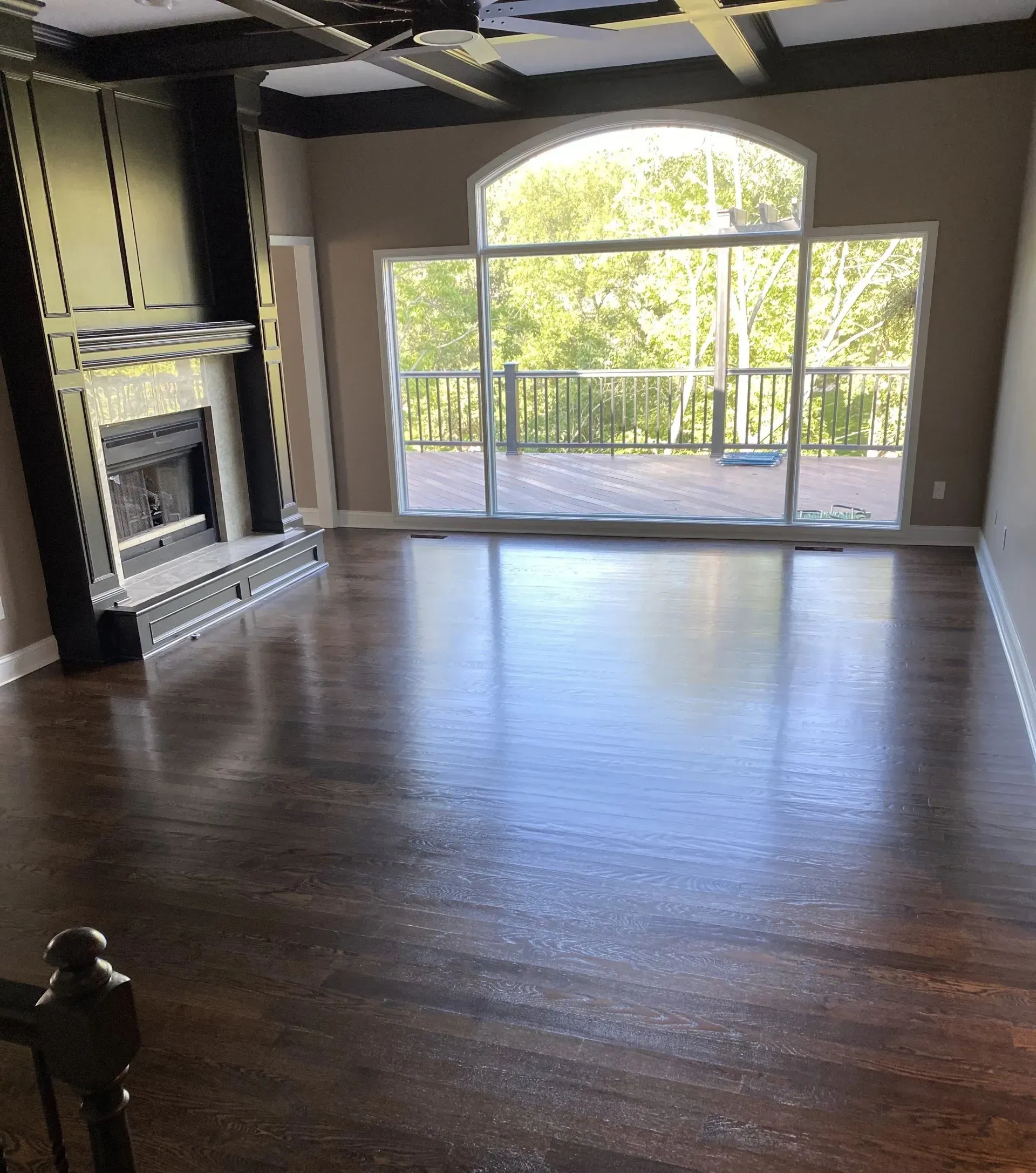 Living room with dark wood floors, fireplace, and large window overlooking a deck and trees.