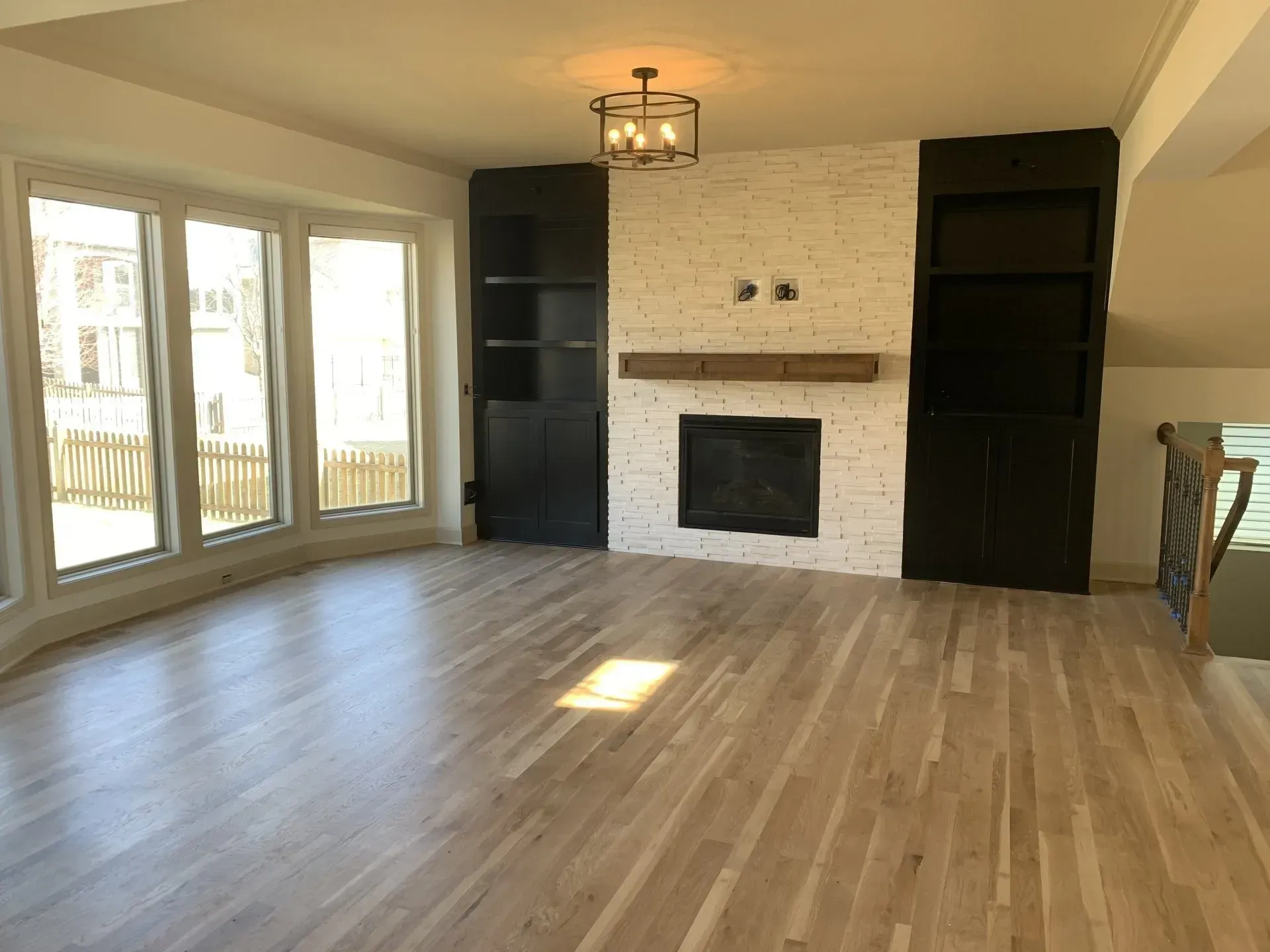 Empty living room with wood flooring, fireplace with white brick, black built-ins, and large windows.