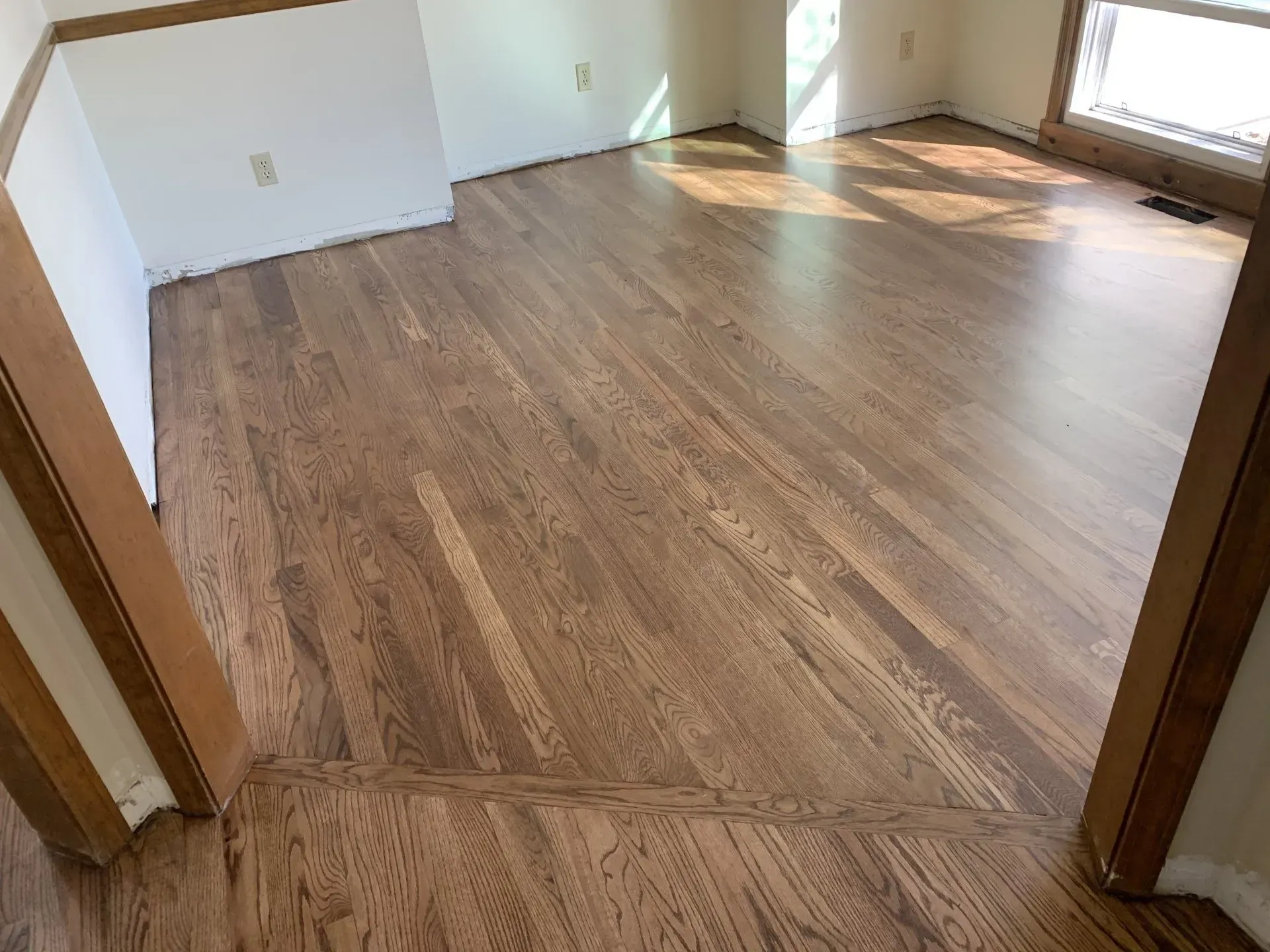 Hardwood floor in a room, with brown wooden planks angled across the view, natural light streaming in from a window.