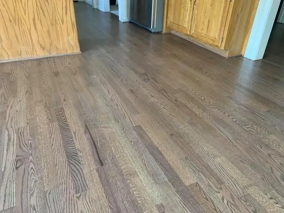 Hardwood floor, brown stained planks with visible wood grain, in a room with a light wood cabinet and white trim.
