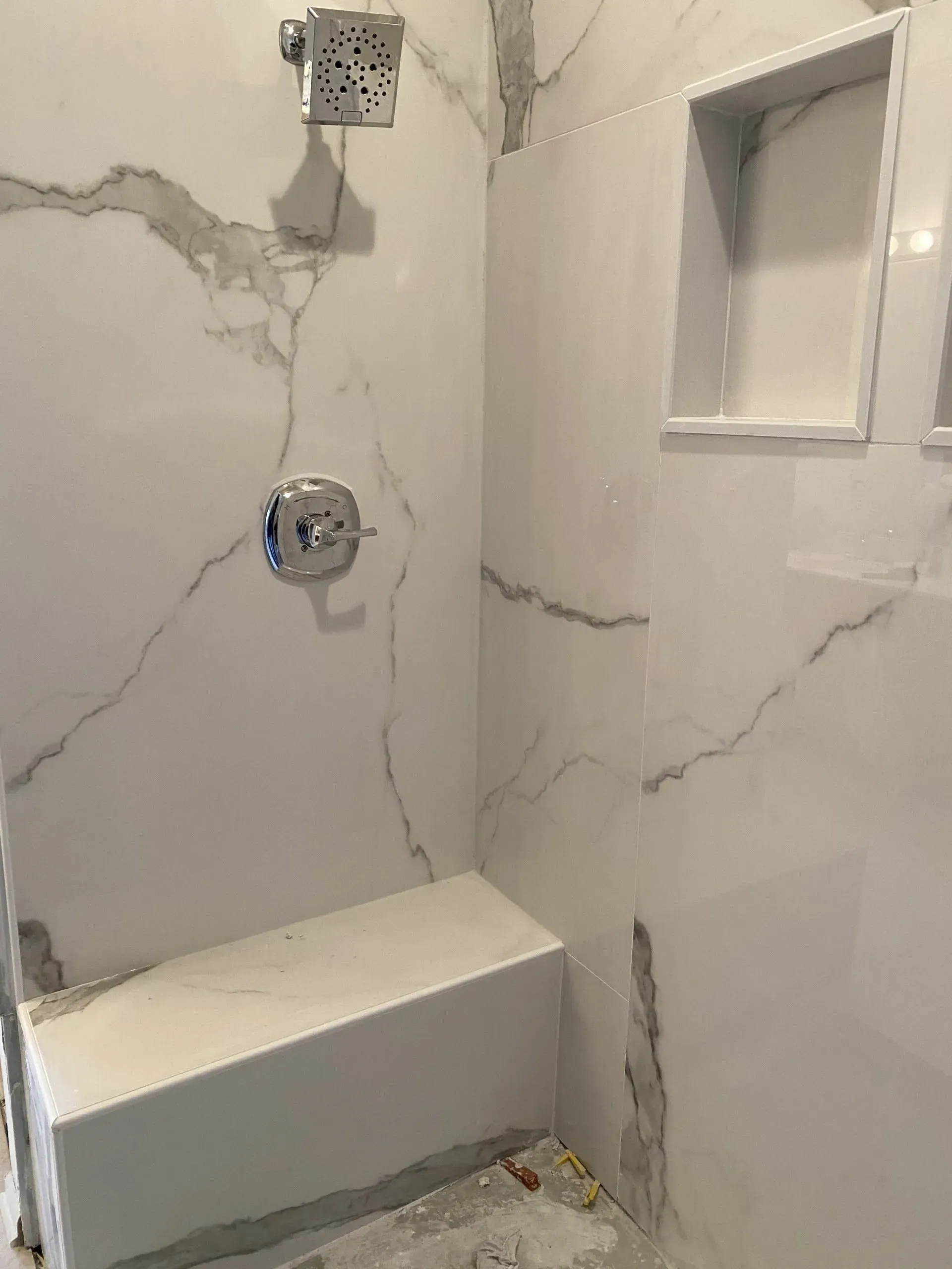 White tiled shower with built-in bench and recessed shelf. Chrome fixtures against marble-like veining.