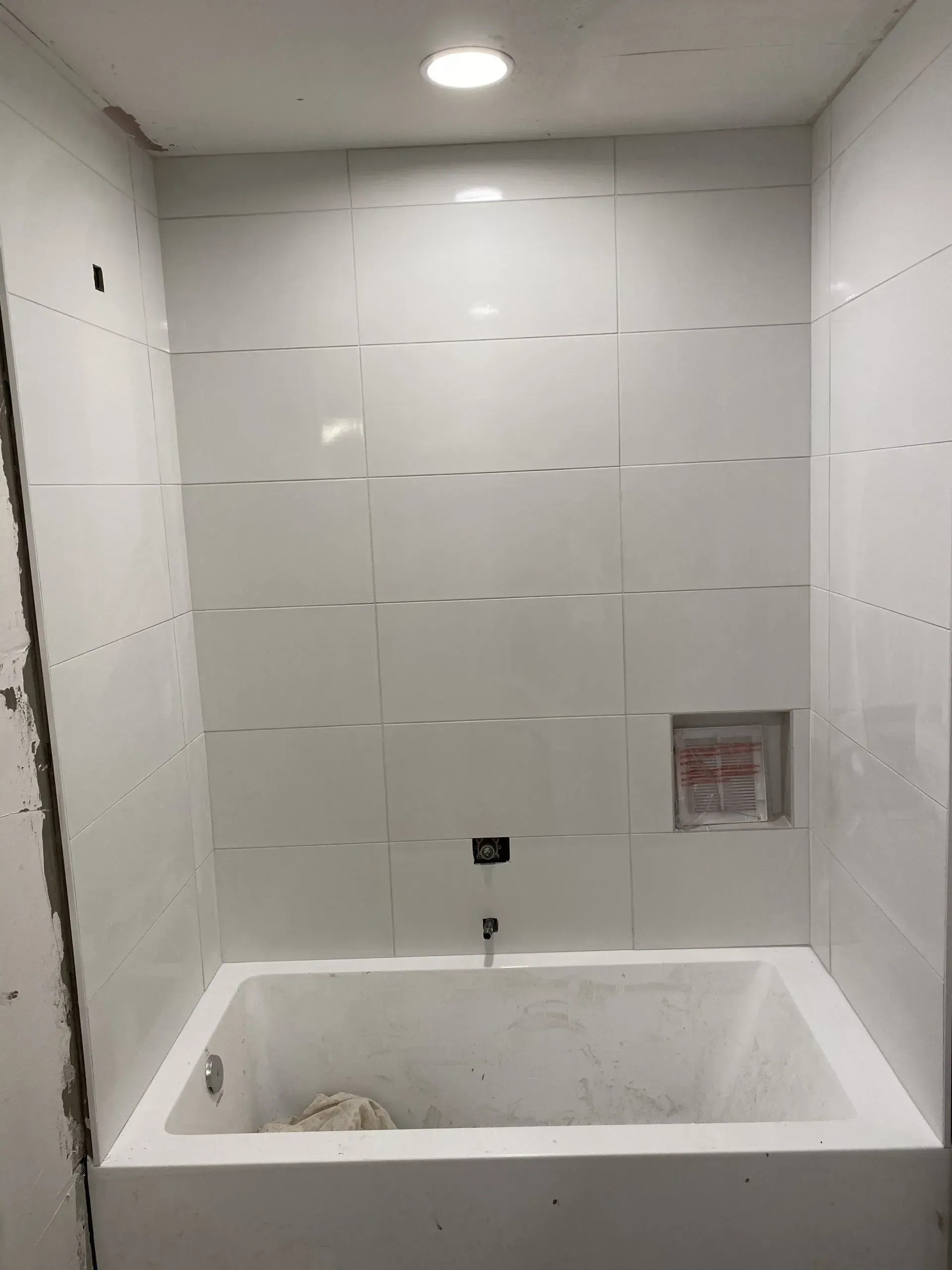 White tiled bathtub and shower in a bathroom with recessed shelf and ceiling light.