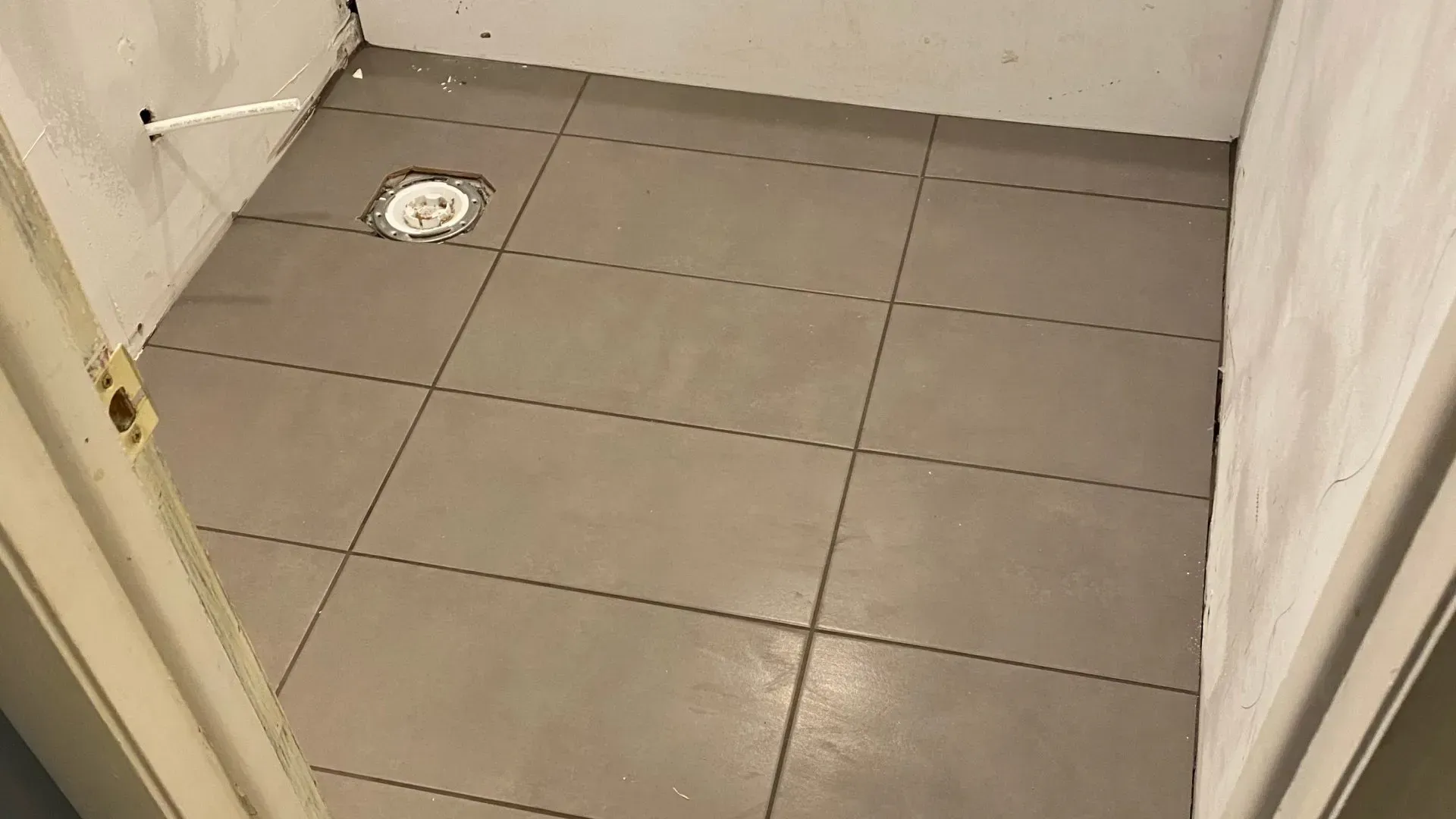 Tiled shower floor in a small room, drain visible. Gray tiles, white walls, viewed from doorway.