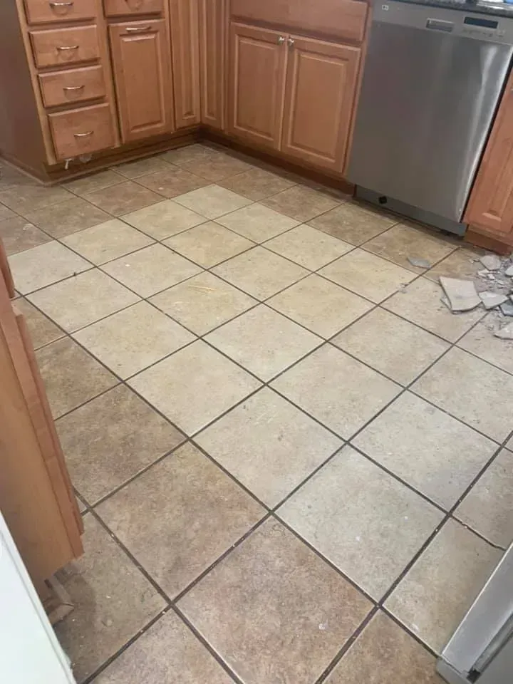 Kitchen floor with tan tiles, some lighter-colored, next to light-brown cabinets and a stainless-steel dishwasher.