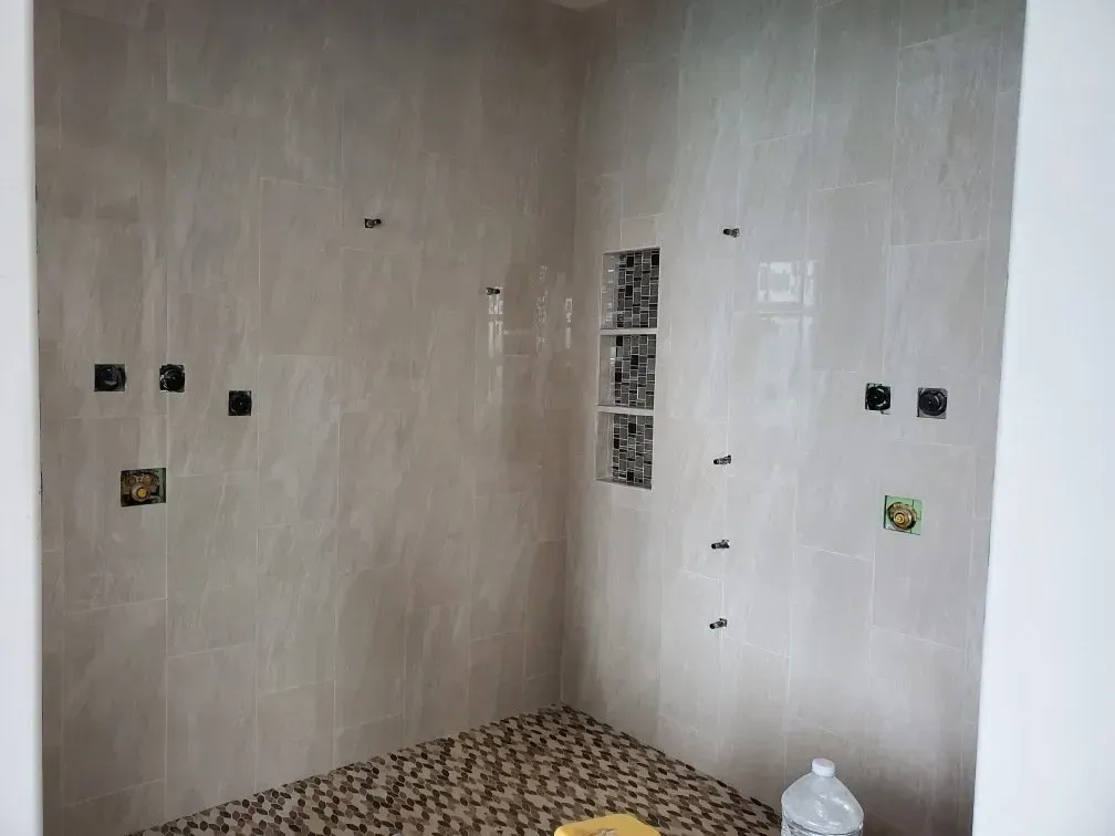 Shower stall with light beige tile walls, pebble floor, and plumbing fixture cutouts.