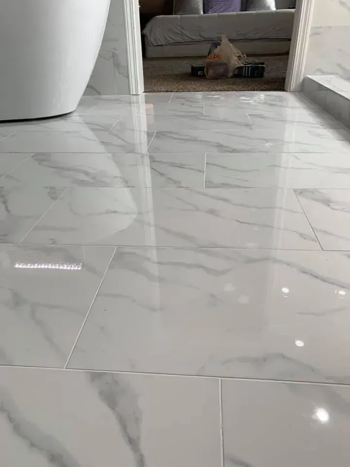 White marble-look tile flooring in a bathroom, reflecting light. A bathtub is visible on the left, doorway to bedroom in the background.