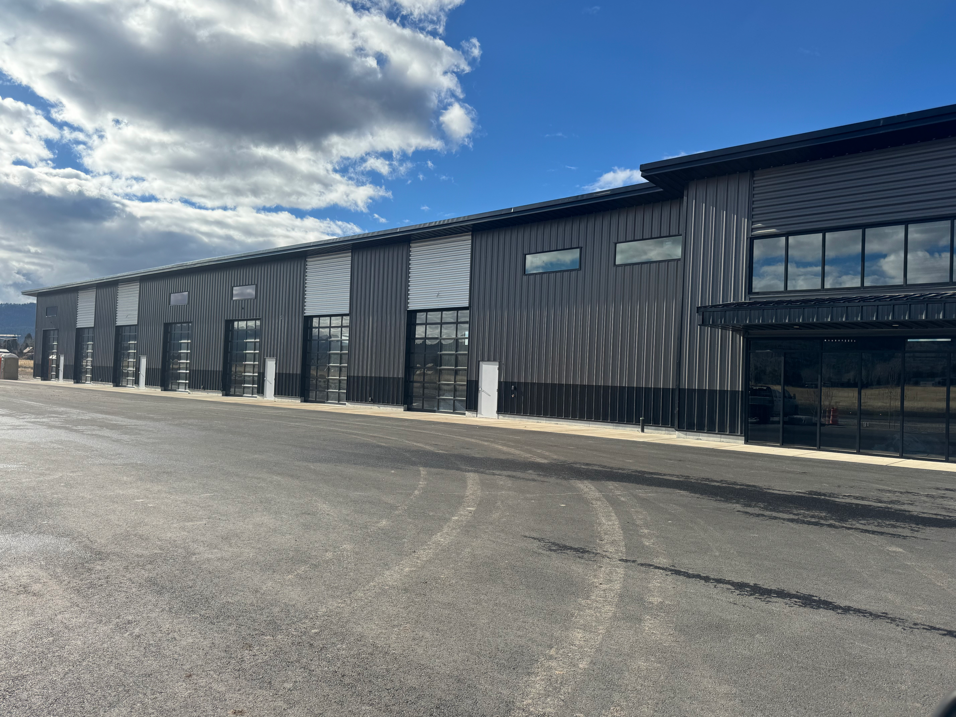 Exterior of a large black industrial warehouse with a wide empty asphalt lot under a partly cloudy sky