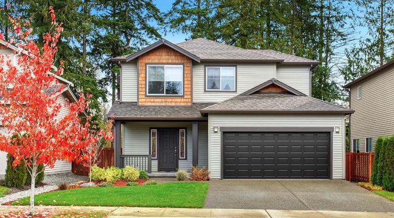 Two-story suburban house with black garage door, gray siding, and a red tree in front yard