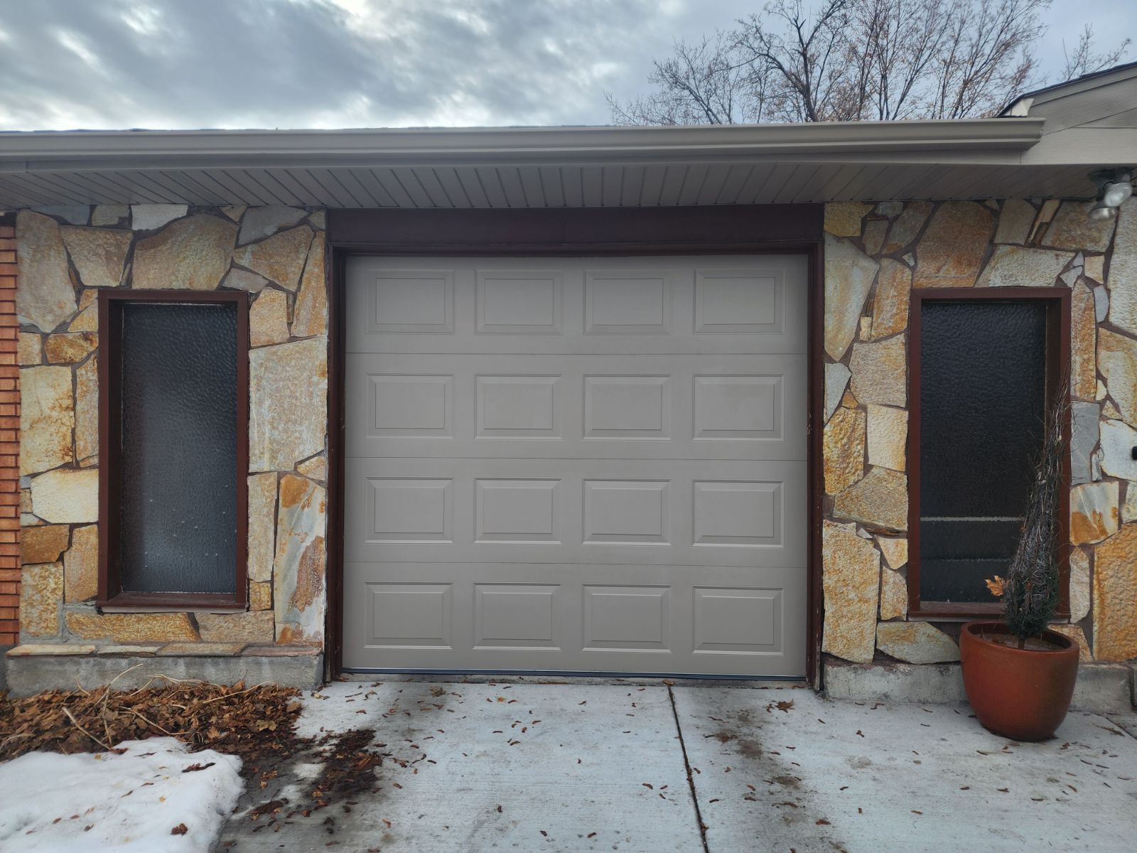 Front entrance with a gray garage door, stone facade, narrow windows, and snow on the driveway