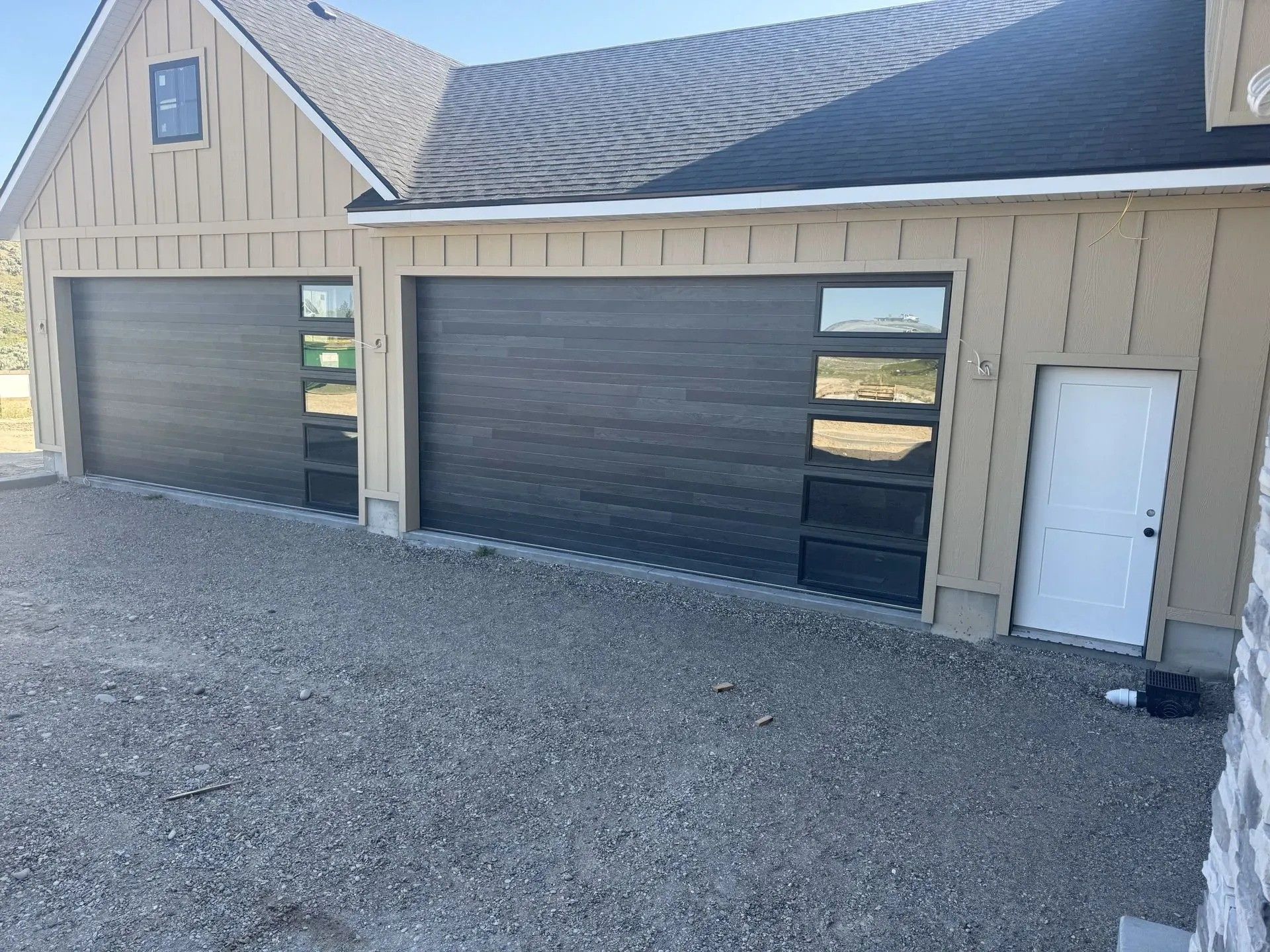 Two-car garage with dark doors, beige siding, and a white side entry door on gravel driveway