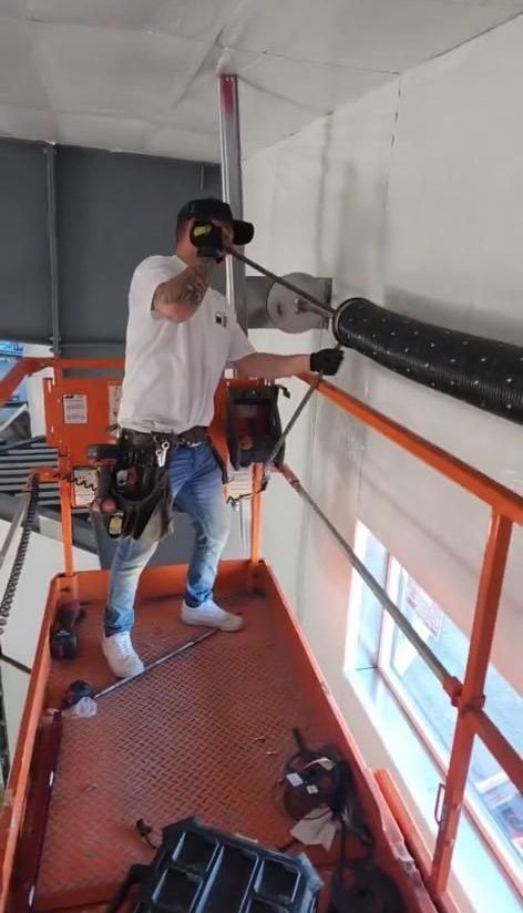 Worker using a headset stands on a scaffold in an unfinished room, inspecting a ceiling pipe.