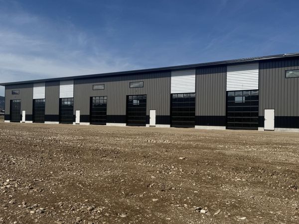 Row of dark gray industrial buildings with black doors on a gravel lot under a clear sky