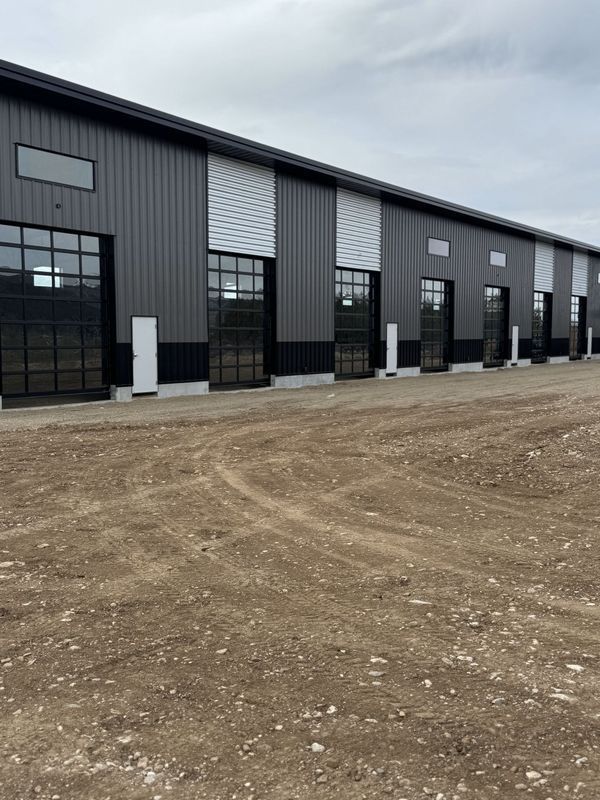 Row of black industrial warehouse units with large windows beside a dirt lot under a gray sky