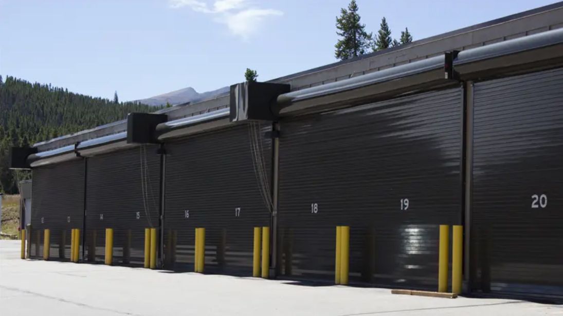 Row of dark garage doors numbered 18, 19, and 20 with yellow bollards in front, snowy mountain backdrop