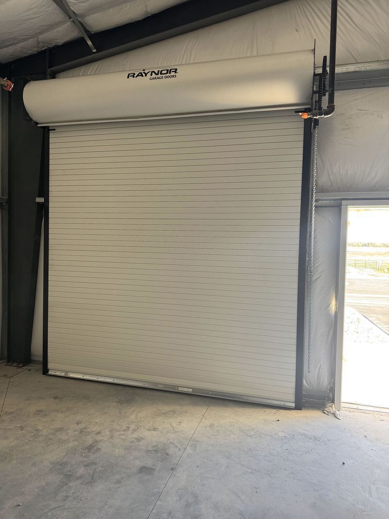 Industrial roll-up door inside a warehouse, partially open with daylight visible outside.