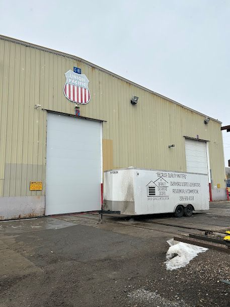 Industrial warehouse exterior with a large garage door and a white dumpster on a wet, snowy lot