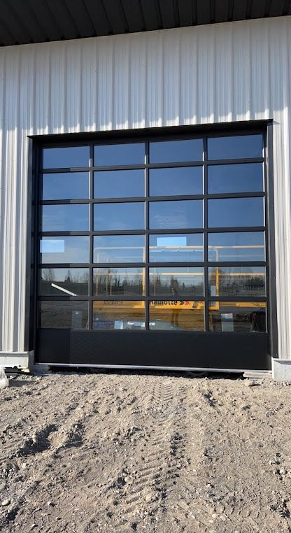 Black garage door with glass panels on a white metal building, set above a gravel ground.