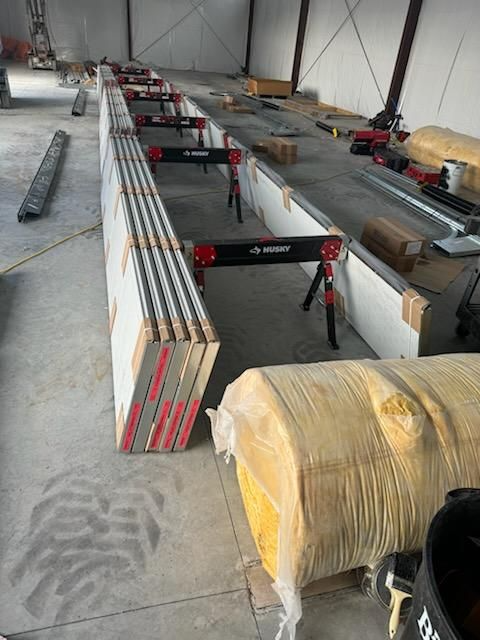 Warehouse aisle with stacked long materials, red sawhorses, and wrapped rolls on a concrete floor.