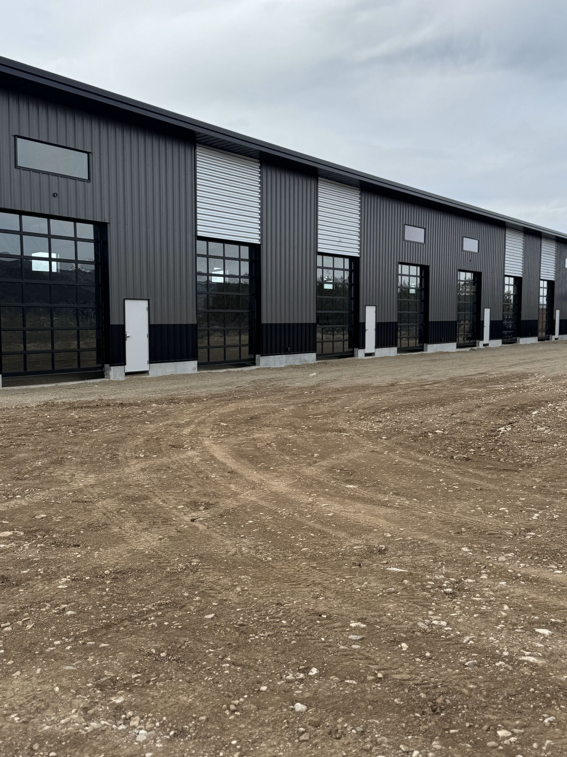 Row of dark industrial warehouse units with large windows beside a gravel lot under a gray sky