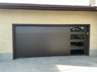 Dark brown modern garage door with horizontal windows on the right side of a brick house.
