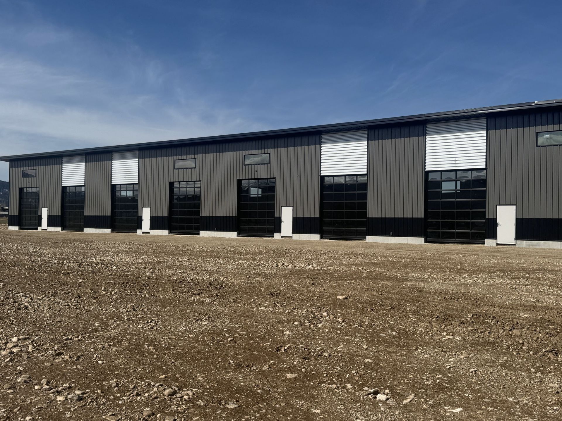 Long gray industrial building with black doors under a clear blue sky, viewed across a gravel lot