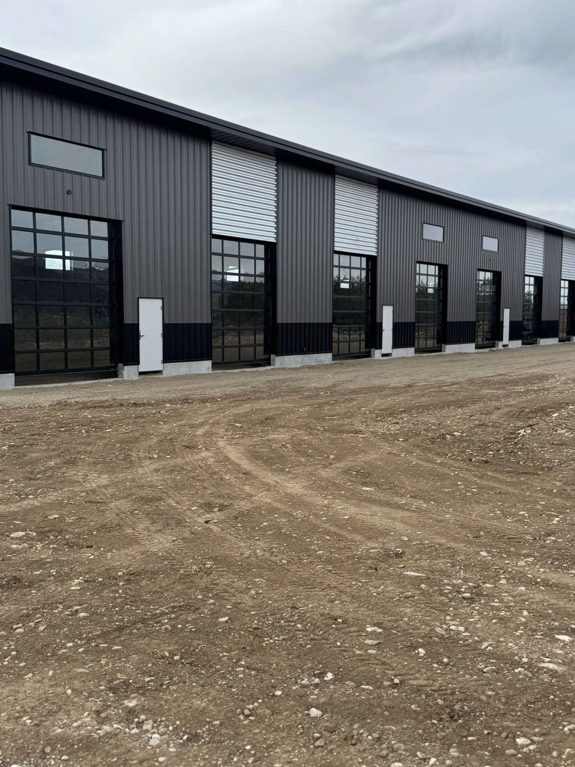 Row of dark gray industrial warehouse units with large windows and gravel lot under cloudy sky