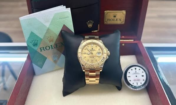 Gold Rolex watch in a box with a green booklet and a small white clock on a table.