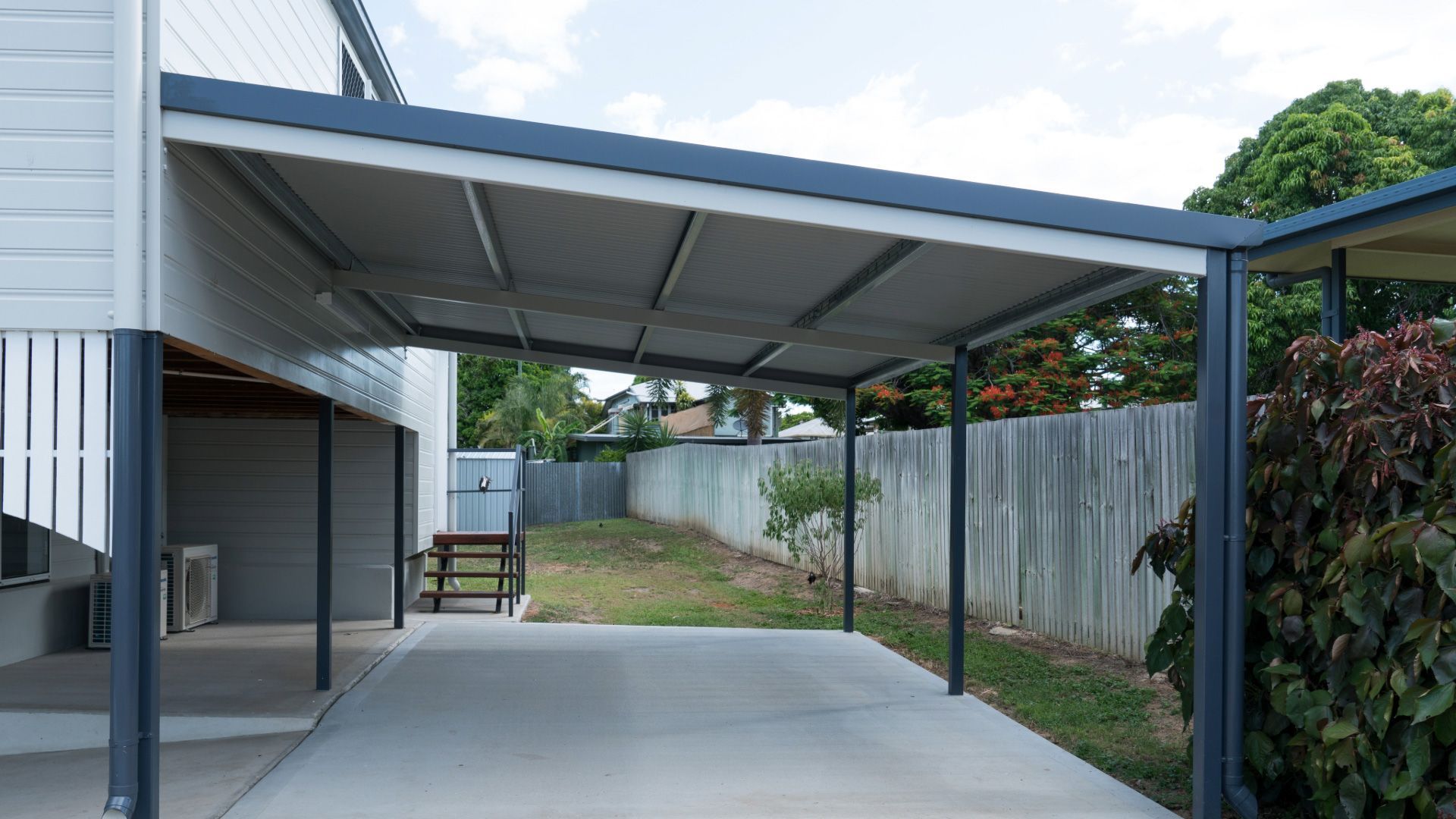 Covered carport beside a house with a concrete driveway and fenced yard under a cloudy sky