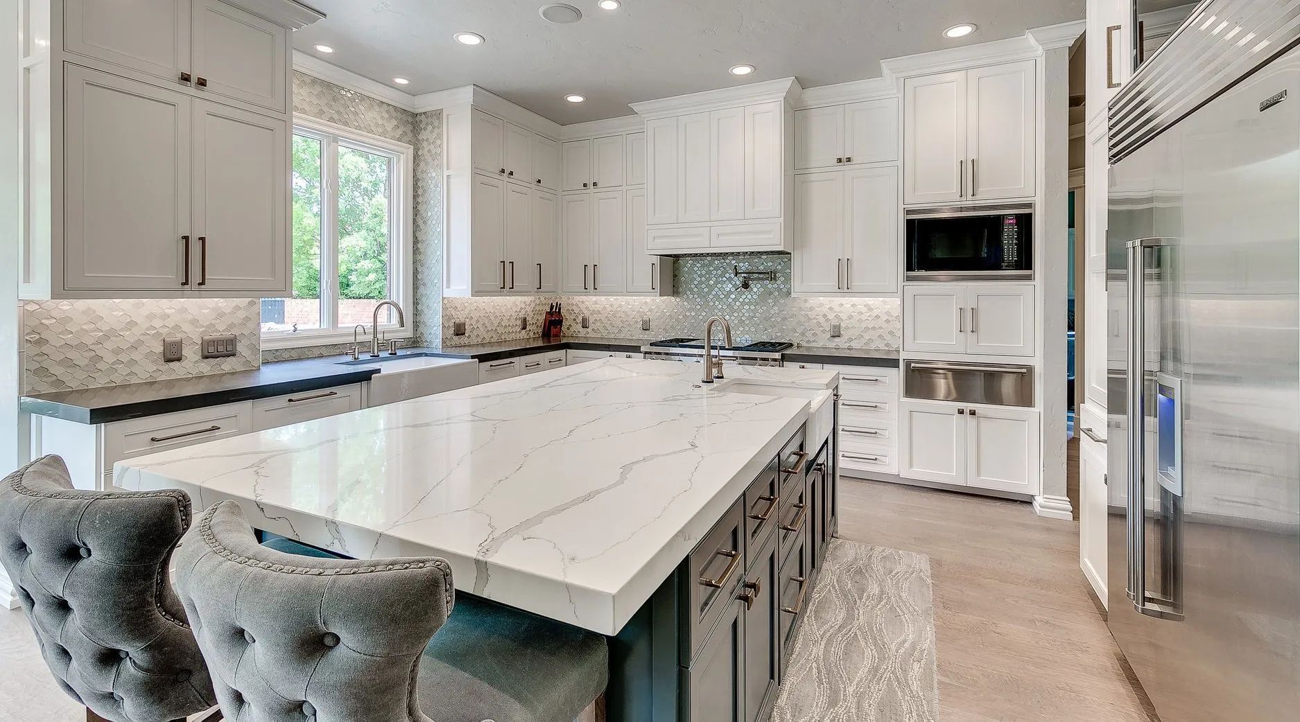 White kitchen with large island, marble countertops, stainless steel appliances, and two upholstered bar stools.